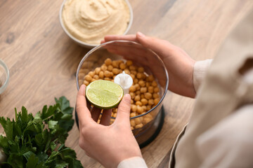 Young woman adding lime juice into blender with chickpeas for hummus on table in kitchen