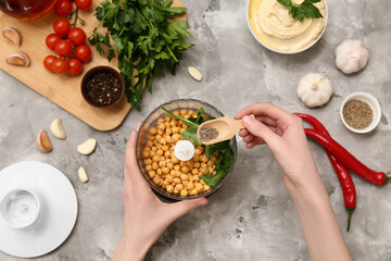 Young woman adding cumin into blender with chickpeas and parsley for hummus on grey grunge...