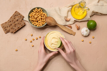 Young woman adding lime juice into bowl of tasty hummus on beige grunge background