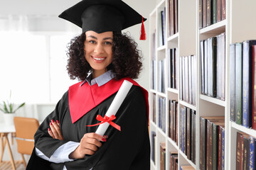 Pretty African-American female graduate with diploma near bookshelves in library