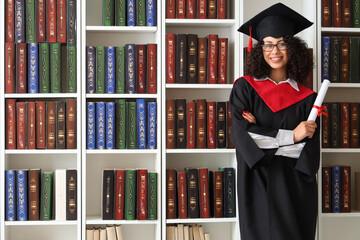 Pretty African-American female graduate in eyeglasses with diploma near bookshelves in library