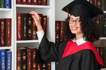 Pretty African-American female graduate in eyeglasses near bookshelves in library