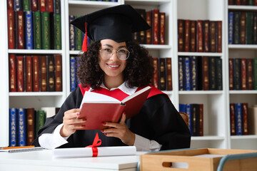 Pretty African-American female graduate with books sitting at table in library