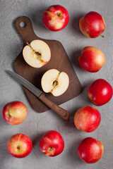 A halved red apple placed on a dark wooden cutting board beside a kitchen knife, surrounded by whole apples on a gray textured surface. Ideal for culinary, recipe, or healthy eating concepts.