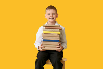 Cute little boy with stack of books sitting on wooden stool against color background