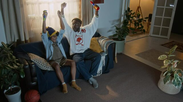 Cheerful African American father and son seated on couch, with snacks and noisemakers in hands, fully engaged in sports game night tradition from comfort of their living room