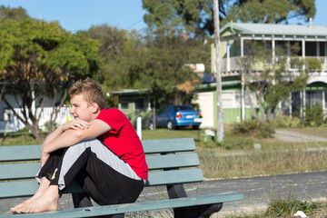 Teenage boy sitting on park bench with arms around knees