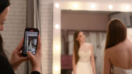 Blogger films bride trying on dress in wedding salon on smartphone, reflection in mirror. Bride and her friend choose wedding dress in boutique, live broadcast. Sales consultant films short video with - Powered by Adobe
