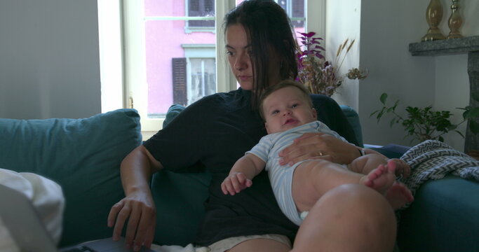 Mother holding her baby while working on a laptop at home, balancing childcare and remote work in a cozy living room setting