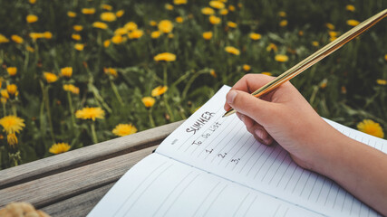 Open notebook with "Summer To Do List" written inside, resting on a wooden bench in a field of blooming dandelions, with a straw hat placed nearby in soft afternoon sunlight