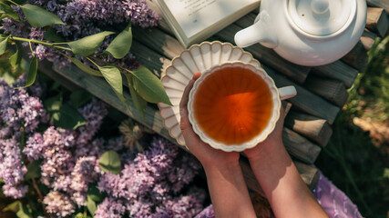 Child holding a teacup on a wooden bench with an open book, teapot, and lilac flowers. Warm...