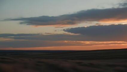 An aerial landscape captures soft pink and gray clouds at sunset, hovering over an expansive, darkened field and distant horizon.