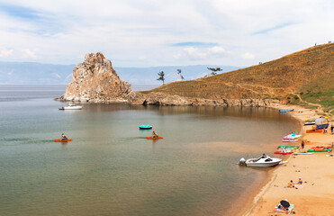 Summer Baikal Lake. Tourists rest near famous Shamanka rock - natural landmark of Olkhon Island, sunbathing on sandy beach, ride water bikes and pleasure boats on sunny August day