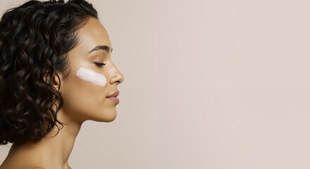 Profile of woman with curly hair and cream product applied to cheek. Eyes closed in relaxed expression shows skincare moment on neutral beige background.