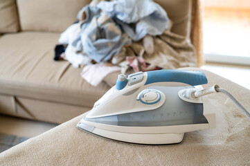 Modern steam iron ready to use, background shows pile of un-ironed laundry in living room