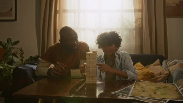 African American father and son playing stacking block game at table in sunlit living room, enjoying time with each other and sharing cozy, family moment