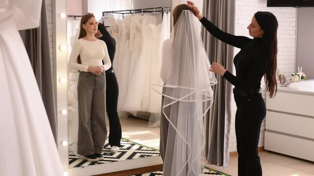 young bride in a wedding salon tries on a dress and veil with the help of a consultant near a large full-length mirror. wedding boutique, planning and preparation for the celebration. Wedding consulta