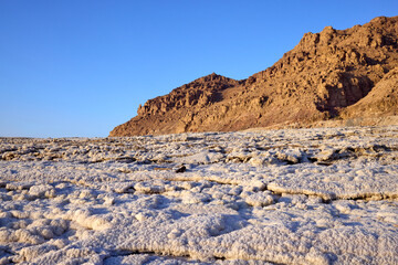 Expansive view of Dead Sea salt flats against mountainous backdrop in Jordan. White crystallized salt plain contrasts with reddish-brown mountains under  blue sky at the world's lowest elevation