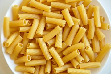 Dry tortiglioni pasta arranged in a white bowl, showing short ridged tubes with a golden yellow hue, ideal for Italian recipes and food backgrounds