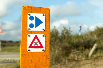 Bright orange warning sign with directional symbols indicating caution and safety measures in a natural outdoor environment with cloudy sky