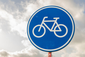 Bicycle lane sign against a cloudy sky, featuring a blue circular design with a white bicycle icon,...