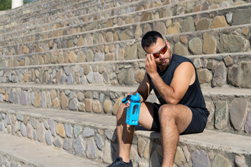 Athletic man sitting on stone steps outdoors resting after workout with sunglasses and water bottle under sunlight