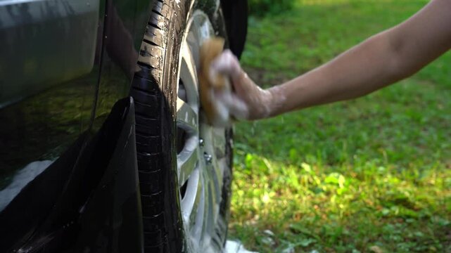 Hands wash car wheel with foam and sponge on lawn with green grass near pond or lake. Concept of washing car in prohibited place in nature
