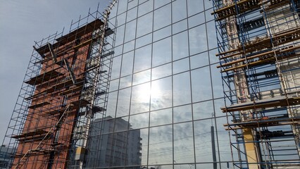 Modern building with reflective glass facade partially covered by scaffolding on both sides, reflecting the sky and surrounding buildings. Concept: urban development and contemporary architecture