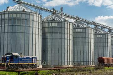 A blue locomotive rests beside a row of towering metal silos in a rural area, showcasing industrial agriculture and machinery © SMK