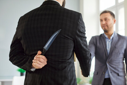 Back view of man in suit hiding knife behind back while shaking hands with another man. Concept of betrayal, deceit, backstabbing or false trust in corporate environment between partners or colleagues