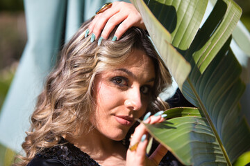 Young blonde woman looks sensually through a giant leaf of a banana tree. The woman has one hand on...