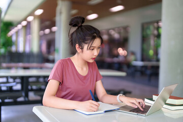 Fototapeta premium Young Asian Woman Studying Online Using Laptop and Writing in Notebook on Campus for Education Project Learning and Exam Preparation