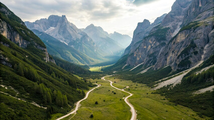 Obraz premium Winding dirt road cuts through lush green valley surrounded by towering mountains under partly cloudy sky, evoking sense of adventure