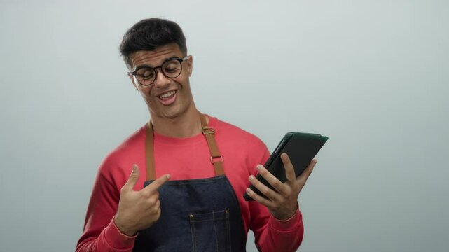 Young man wearing glasses and apron smiles while holding tablet and giving thumbs up against white background