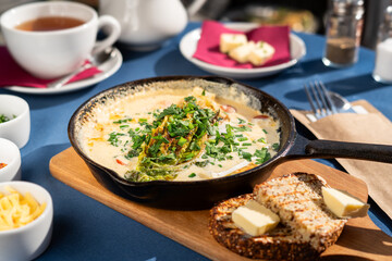 Baked scrambled eggs with goat cheese and vegetables served in a cast iron skillet with dark whole wheat bread, chives, yellow cheese and greens. A breakfast set, illuminated by the morning sun.