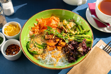 Rice noodles served with tofu, asparagus, avocado chunks, red lettuce and roasted peanut pieces. Served in a green bowl. A breakfast set illuminated by the morning sun.