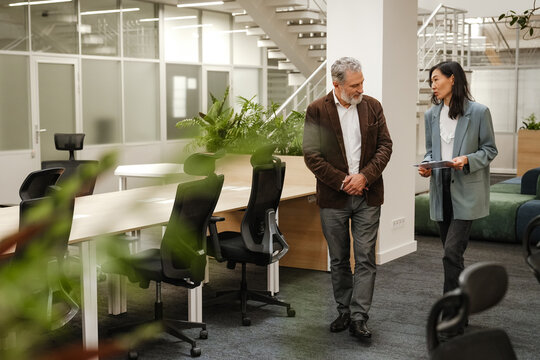 A middle-aged man and woman are walking and discussing documents in a modern open-plan office. They are business partners engaged in a professional conversation near workstations.