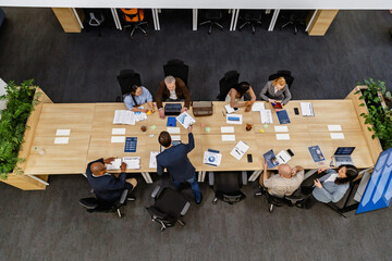 A diverse group of adult men and women collaborate around a large table during a business meeting...