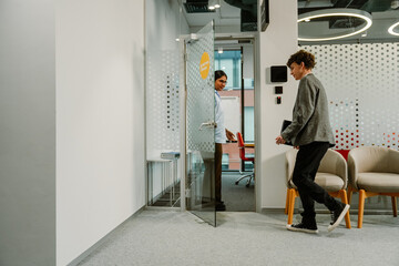 An adult Indian woman holding the door open for a curly-haired adult man entering a modern office meeting room. The space features glass walls, a light interior, and casual business attire.