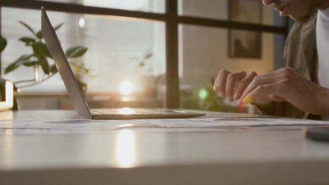 Young businessman summarizing statistical data while looking through financial papers and using laptop at workplace