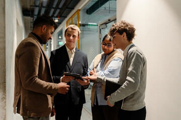 A group of diverse adult coworkers, including Asian and White professionals, stand in a hallway, discussing a project using a tablet during a collaborative work conversation.