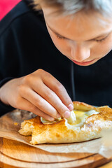 Boy eating traditional khachapuri bread with cheese and egg filling on wooden board close-up view, concept of traditional cuisine enjoyment