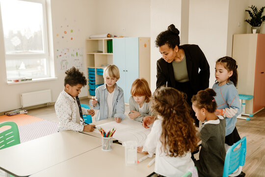 A diverse group of school-aged students collaborates on a large drawing project in a classroom, guided by a teacher. They focus on showcasing teamwork and creativity. - Powered by Adobe