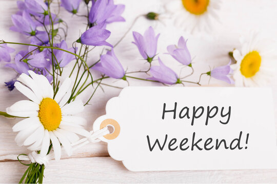 Happy weekend message on white gift tag with daisies and violet bellflowers on rustic wooden background, floral greeting card design, top view composition for summer celebration or weekend wishes