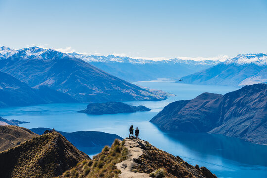 This stunning panoramic landscape captures two hikers standing at the viewpoint on Roy's Peak Track, overlooking the crystal-blue waters of Lake Wanaka on New Zealand's South Island.