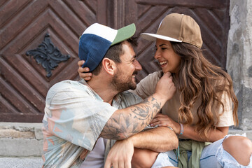 Young happy hipster millennial gen z couple in love and modern clothes sitting outdoor on the street talking and having fun. Man and woman university students meet at college started relationship.