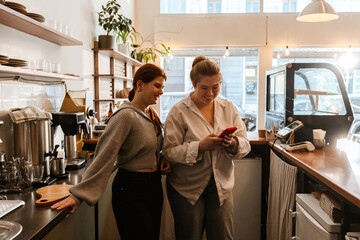 Naklejka premium Two adult White women stand behind the counter in a modern cafe. One woman shows something on her red smartphone while they smile, chat, and work together during the setup.