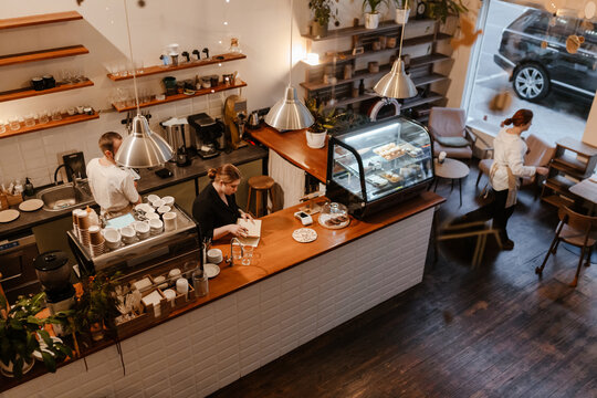 A group of white adults, including baristas and waitstaff, prepare coffee and serve tables at a cozy cafe with wooden floors and a pastry display counter. - Powered by Adobe