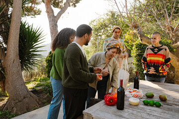 A group of young multiracial men and women stand outdoors around a wooden table with wine, fruits, and vegetables, preparing food together.