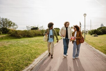 A multiracial group of adult men and a woman walking and talking in a sunny city park, casually dressed and carrying a paper bag, camera, and volleyball on a paved path surrounded by greenery.